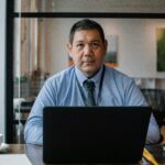 Confident ethnic man wearing formal clothes and tie working at netbook while sitting at table in workspace